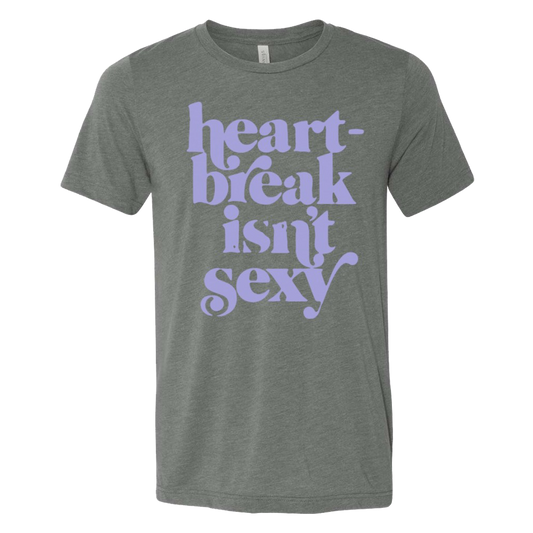 A gray unisex crew neck t-shirt with the phrase 'heart-break isn't sexy' printed in purple ink on the front.